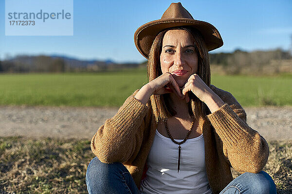 Beautiful woman with hand on chin sitting on land against sky