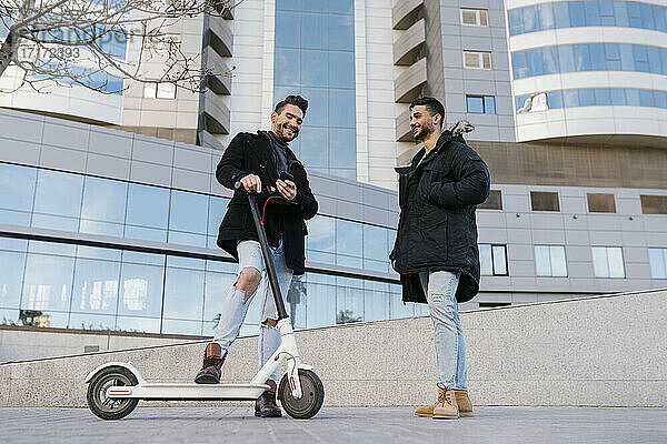 Young man smiling at male friend with electric push scooter while standing against buildings in city
