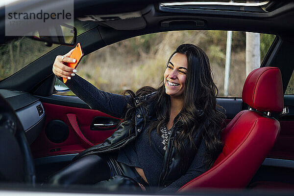 Lächelnde Frau  die ein Selfie mit ihrem Smartphone macht  während sie im Auto sitzt