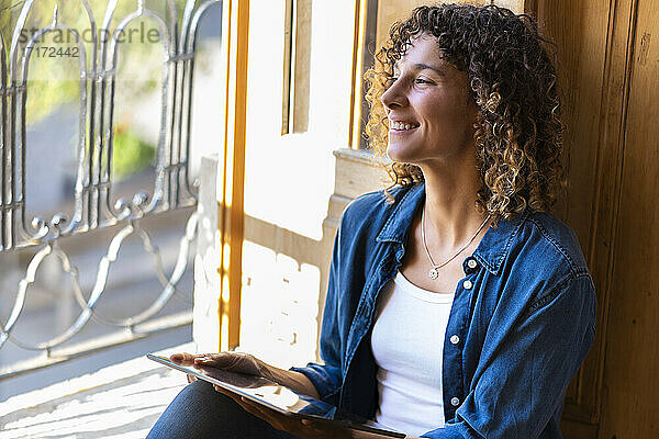 Young woman smiling looking out of window while holding digital tablet at home