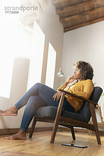 Nachdenkliche Frau mit Hand am Kinn  die wegschaut  während sie zu Hause auf einem Sessel sitzt