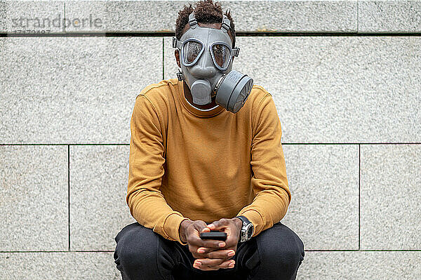 Young man wearing pollution gas mask sitting with mobile phone against wall