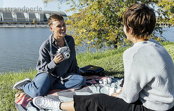 Lächelnde Frau mit Kamera  die einen Freund ansieht  während sie im Park am Fluss sitzt