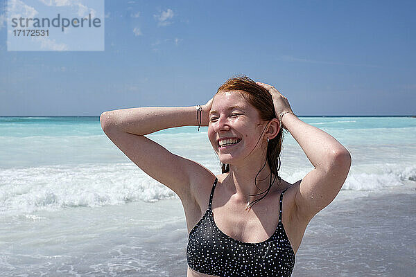 Fröhliche junge Frau steht mit Hand in den Haaren am Strand während eines sonnigen Tages