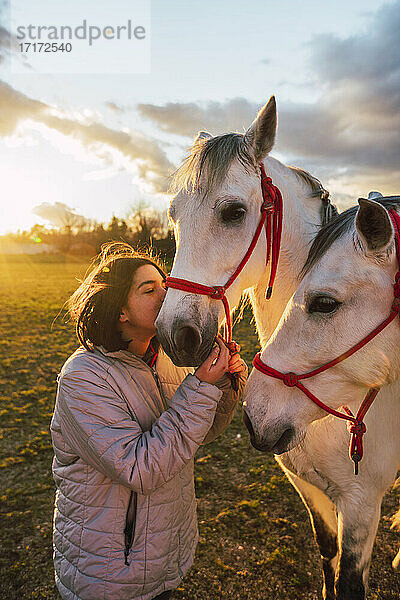 Girl wearing jacket kissing horses while standing in ranch