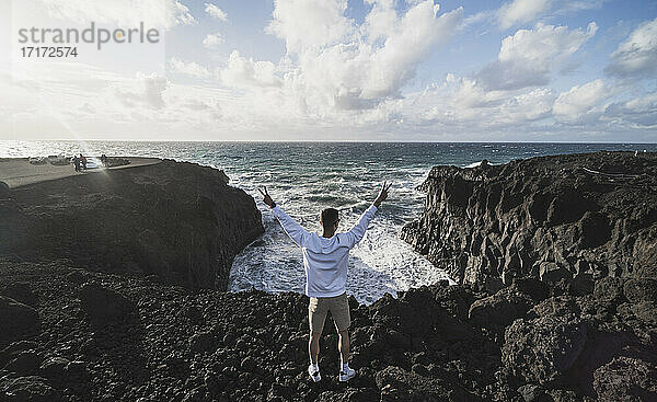 Männlicher Tourist macht ein Friedenszeichen  während er an einem sonnigen Tag in Los Hervideros  Lanzarote  Spanien  auf einem Vulkanfelsen steht.