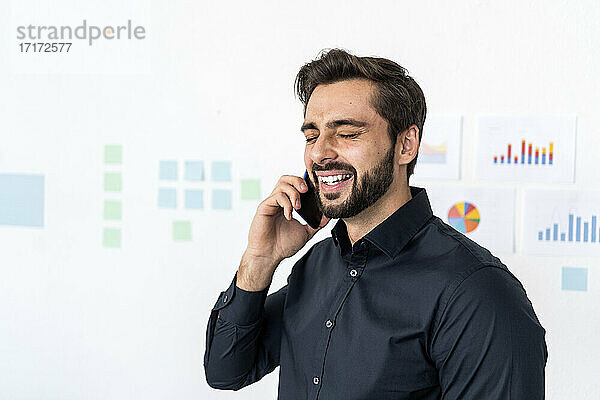 Smiling businessman talking on smart phone with eyes closed against wall in office