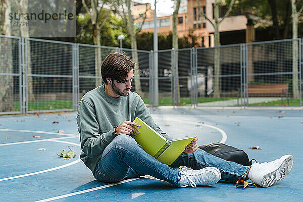 Junger Mann studiert beim Lesen eines Buches auf einem Basketballplatz in der Universität