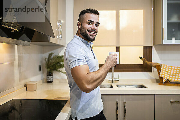 Lächelnder muskulöser Mann  der eine Kaffeetasse hält  während er sich zu Hause an den Küchentisch lehnt