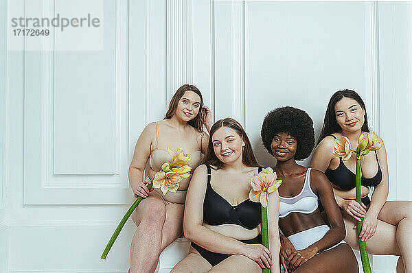 Lächelnde multiethnische Gruppe weiblicher Modelle in Unterwäsche  die Kunstblumen vor einer weißen Wand halten