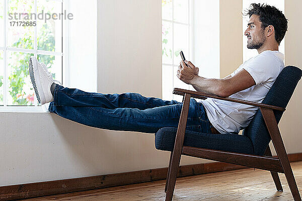 Mann mit Smartphone schaut durch das Fenster  während er sich zu Hause auf einem Sessel entspannt