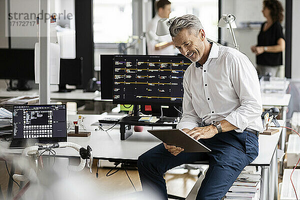 Lächelnder Geschäftsmann  der ein digitales Tablet benutzt  während er auf dem Schreibtisch sitzt  mit Kollegen im Hintergrund in einem Großraumbüro