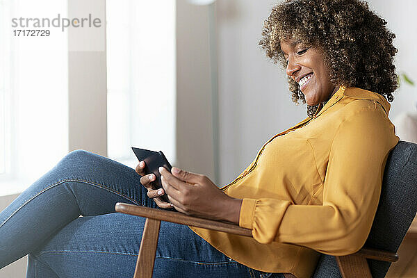 Frau lächelt  während sie ein digitales Tablet benutzt und sich auf einem Sessel zu Hause entspannt