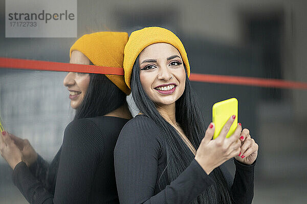 Smiling young beautiful woman with smart phone against glass