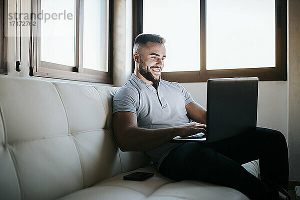 Lächelnder Geschäftsmann arbeitet am Laptop  während er zu Hause auf dem Sofa sitzt