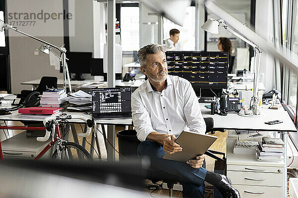 Geschäftsmann mit digitalem Tablet  der wegschaut  während er in einem Großraumbüro mit Kollegen im Hintergrund sitzt