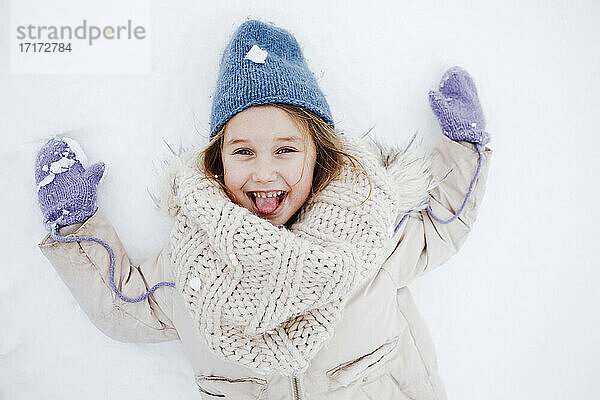 Fröhliches Mädchen mit herausgestreckter Zunge auf Schnee liegend in den Ferien