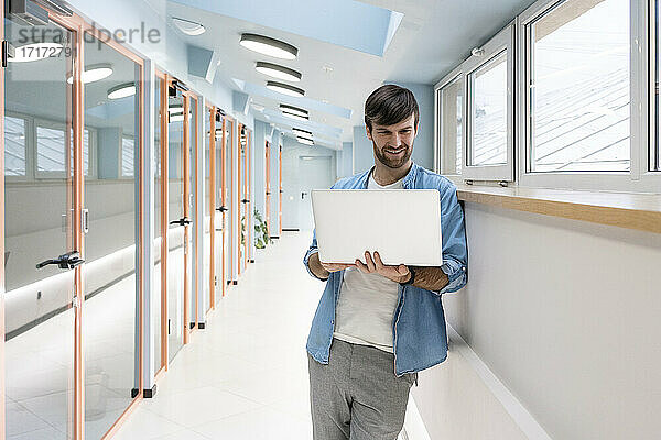 Kreativer männlicher Unternehmer  der an einem Laptop arbeitet  während er sich an die Wand im Korridor lehnt
