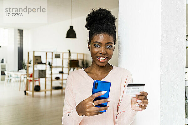 Junge Frau mit Kreditkarte  die lächelnd über ihr Handy einkauft  während sie zu Hause steht
