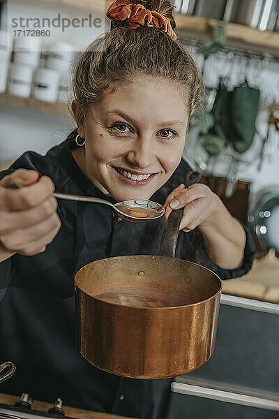 Smiling chef tasting broth soup in saucepan while standing in kitchen