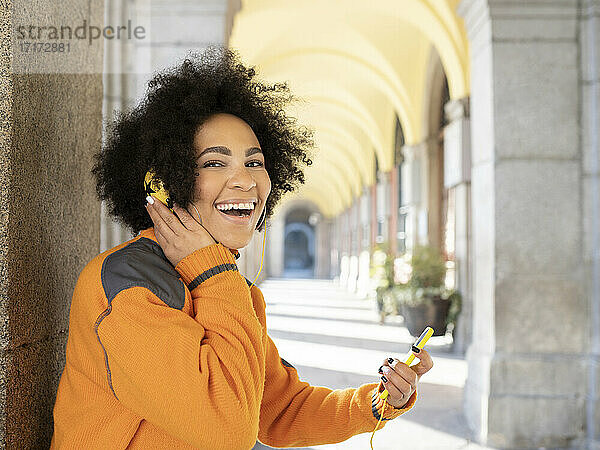Cheerful woman listening music while using mobile phone standing against pillar