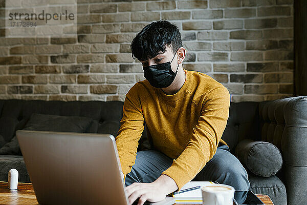 Mann mit Gesichtsschutz  der während der COVID-19 in einem Café sitzt und einen Laptop benutzt