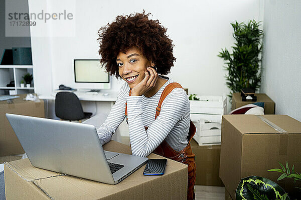 Lächelnde junge Frau mit Hand am Kinn vor einem Laptop im Wohnzimmer ihres neuen Hauses