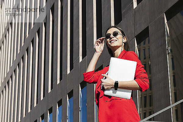 Smiling businesswoman with sunglasses holding laptop while standing against building
