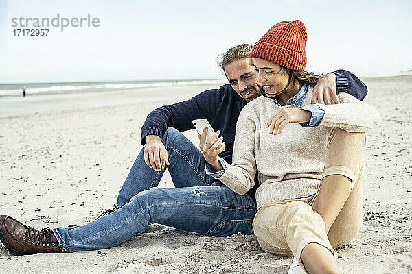 Junges Paar sitzt zusammen am Strand und benutzt ein Smartphone