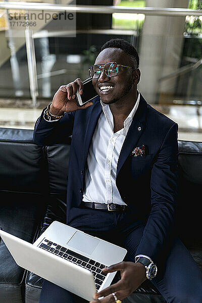 Lächelnder junger männlicher Unternehmer  der in der Hotellobby mit einem Laptop auf einem Smartphone spricht