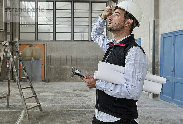 Männlicher Architekt mit digitalem Tablet und Bauplänen  der in einem Gebäude steht und nach oben schaut