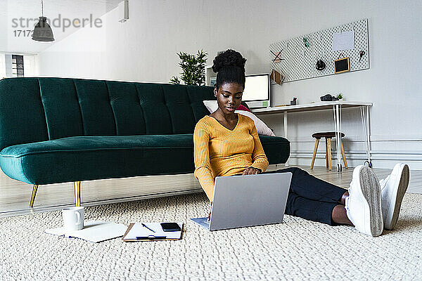 Young Afro woman using laptop while sitting on carpet against sofa in living room at home