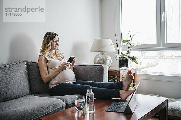 Schwangere Frau benutzt ihr Smartphone  während sie zu Hause auf dem Sofa sitzt