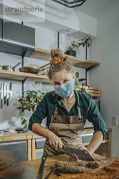 Young female chef wearing protective face mask cutting salami while working in kitchen during COVID-19