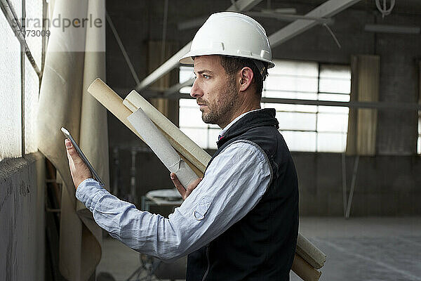 Männlicher Immobilienentwickler  der einen Bauplan mit einem digitalen Tablet hält  während er in einem Gebäude steht