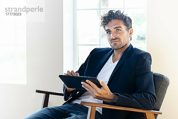 Nachdenklicher Geschäftsmann mit digitalem Tablet  der wegschaut  während er zu Hause auf einem Sessel sitzt