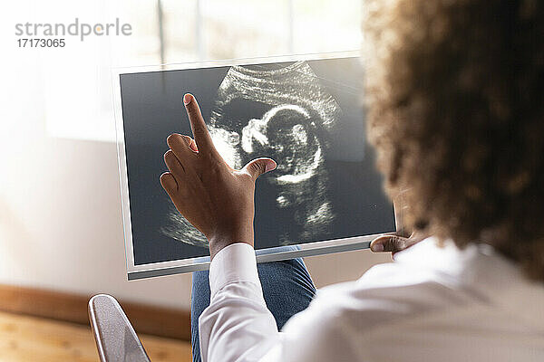 Frau betrachtet Ultraschallbild des Babys auf transparentem Bildschirm  während sie zu Hause sitzt