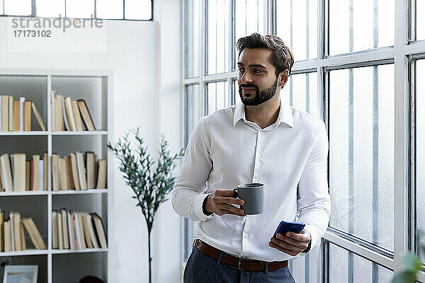Lächelnder Geschäftsmann mit Mobiltelefon  der eine Kaffeetasse hält  während er im Büro wegschaut