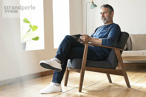 Älterer Geschäftsmann mit digitalem Tablet  der wegschaut  während er zu Hause auf einem Sessel sitzt