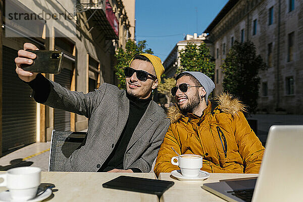 Junge schwule Männer mit Sonnenbrille  die ein Selfie mit ihrem Handy machen  während sie in einem Straßencafé sitzen