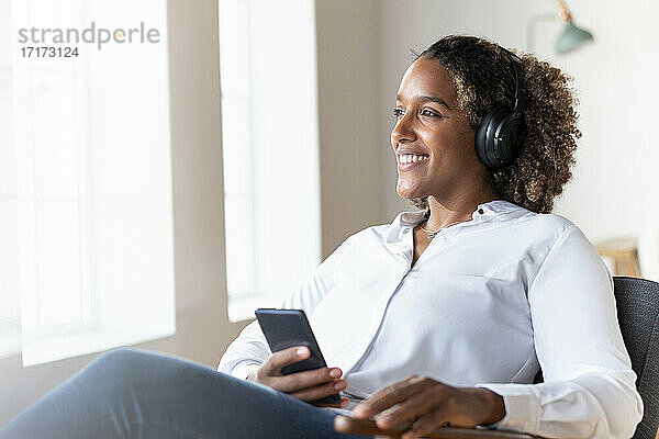 Frau mit Kopfhörern  die wegschaut  während sie mit ihrem Mobiltelefon auf einem Sessel zu Hause sitzt