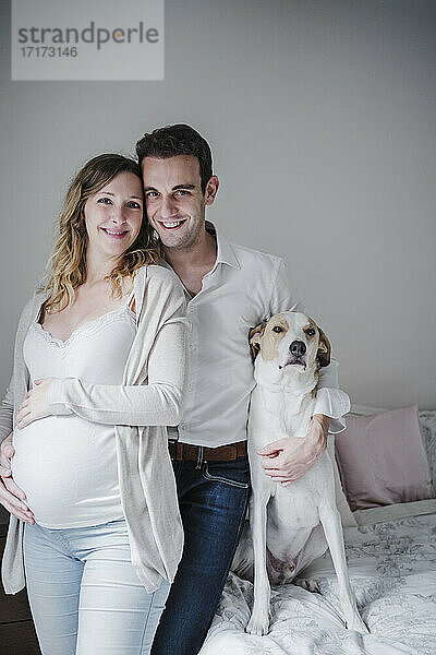 Lächelnder Mann mit schwangerer Frau und Hund im Schlafzimmer stehend