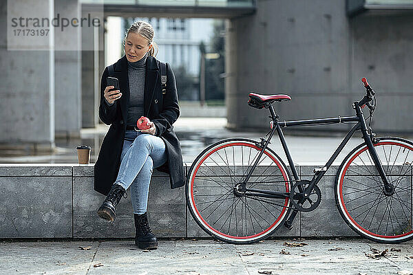 Young woman using mobile phone while sitting on retaining wall by bicycle