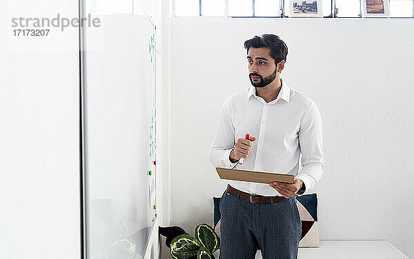 Männlicher Unternehmer mit Filzstift und Klemmbrett  der bei der Arbeit im Büro auf ein Whiteboard schaut