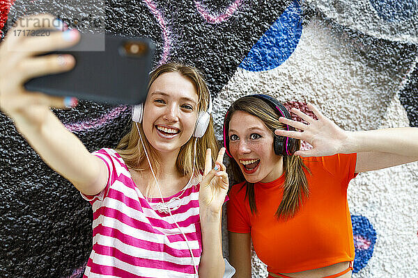 Glückliche Freundinnen machen ein Selfie mit ihrem Smartphone  während sie gegen eine Graffiti-Wand gestikulieren