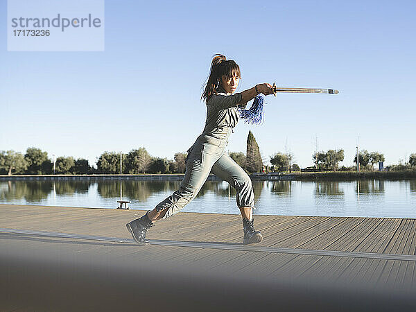 Weibliche Athletin übt Kampfsport mit Schwert am See gegen den klaren Himmel