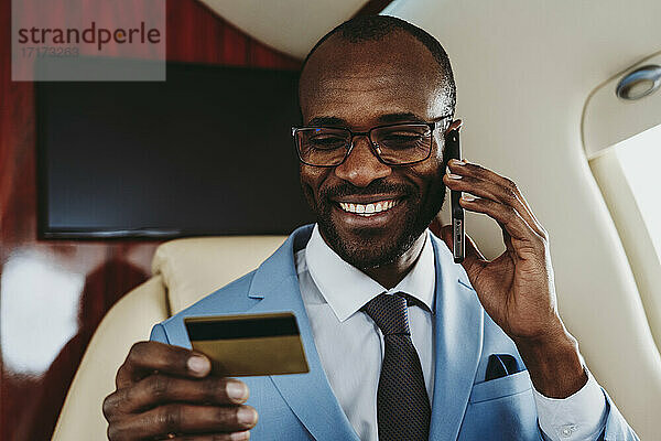 Lächelnder Geschäftsmann  der eine Kreditkarte in der Hand hält  während er im Flugzeug mit seinem Handy telefoniert