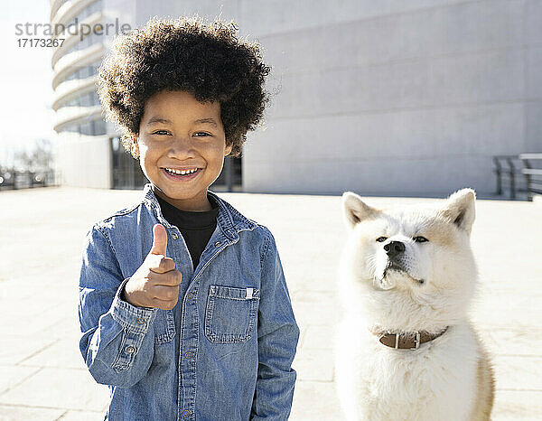 Lächelnder Junge  der neben einem auf dem Gehweg sitzenden Hund steht und den Daumen hochstreckt