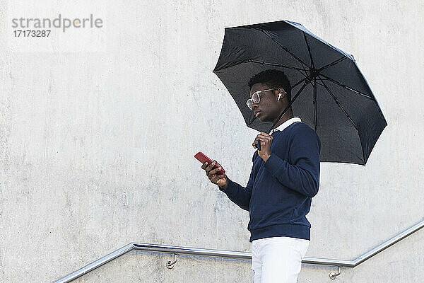 Young African man with umbrella listening music through in ear headphones while using smart phone