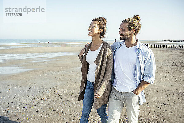Lächelnde Freundin und Freund schauen weg  während sie am Wochenende am Strand spazieren gehen